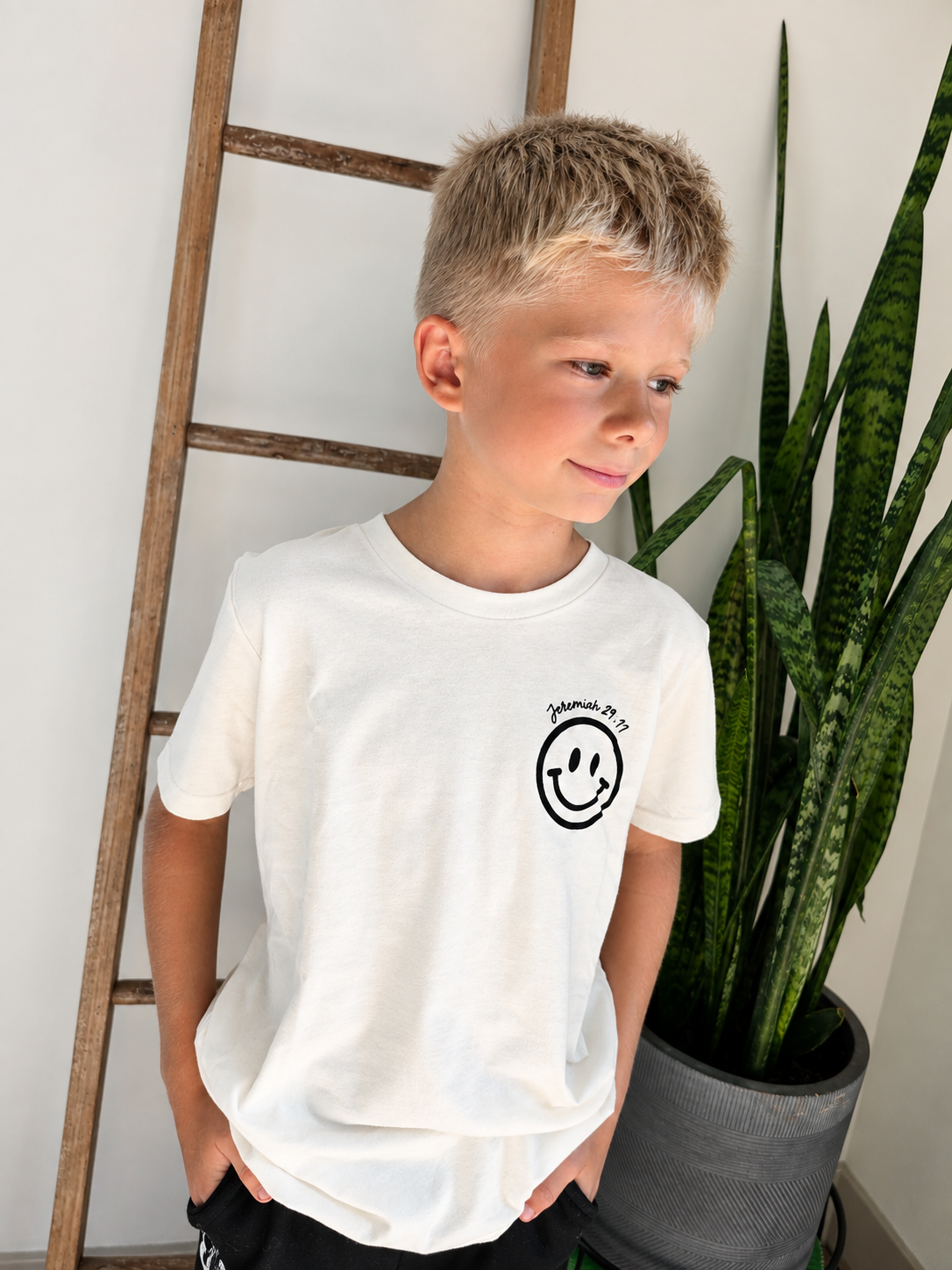 A young boy wearing a white short-sleeve t-shirt with a black smiley face graphic on the left chest, standing indoors next to a wooden ladder and a potted snake plant.