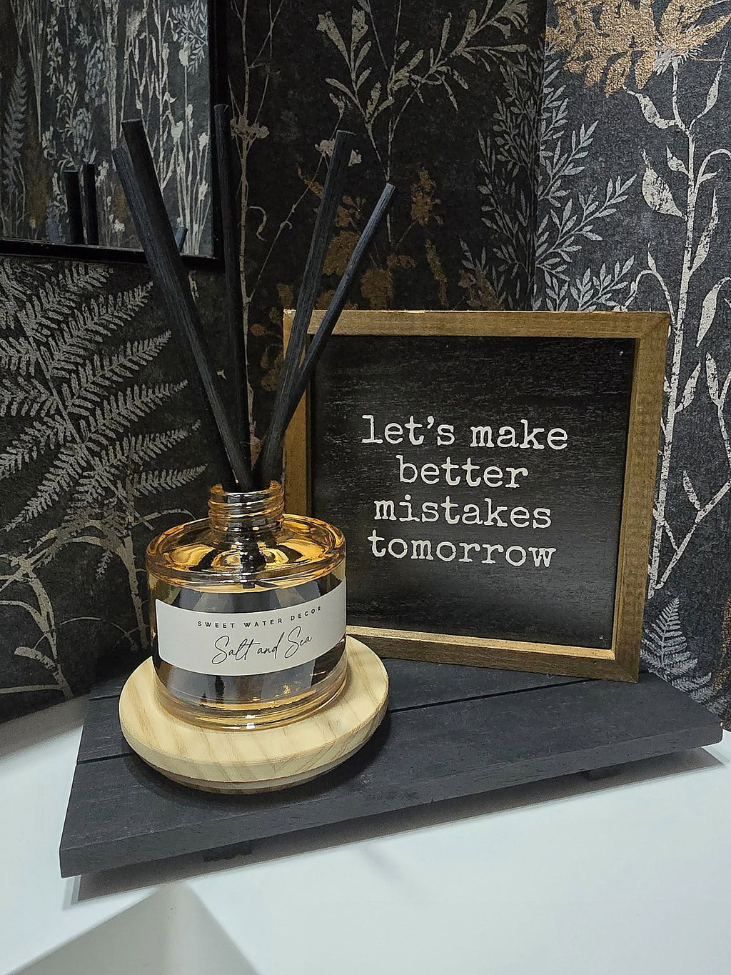 A reed diffuser bottle with black reeds sits on a round wooden coaster next to a small framed sign that reads 'let's make better mistakes tomorrow'. The scene is styled against dark botanical wallpaper.