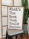 A framed typographic wall sign reading “What’s The Best That Could Happen” in black text on an off-white background, styled leaning on a stool in a home setting with a plant and wooden ladder visible.