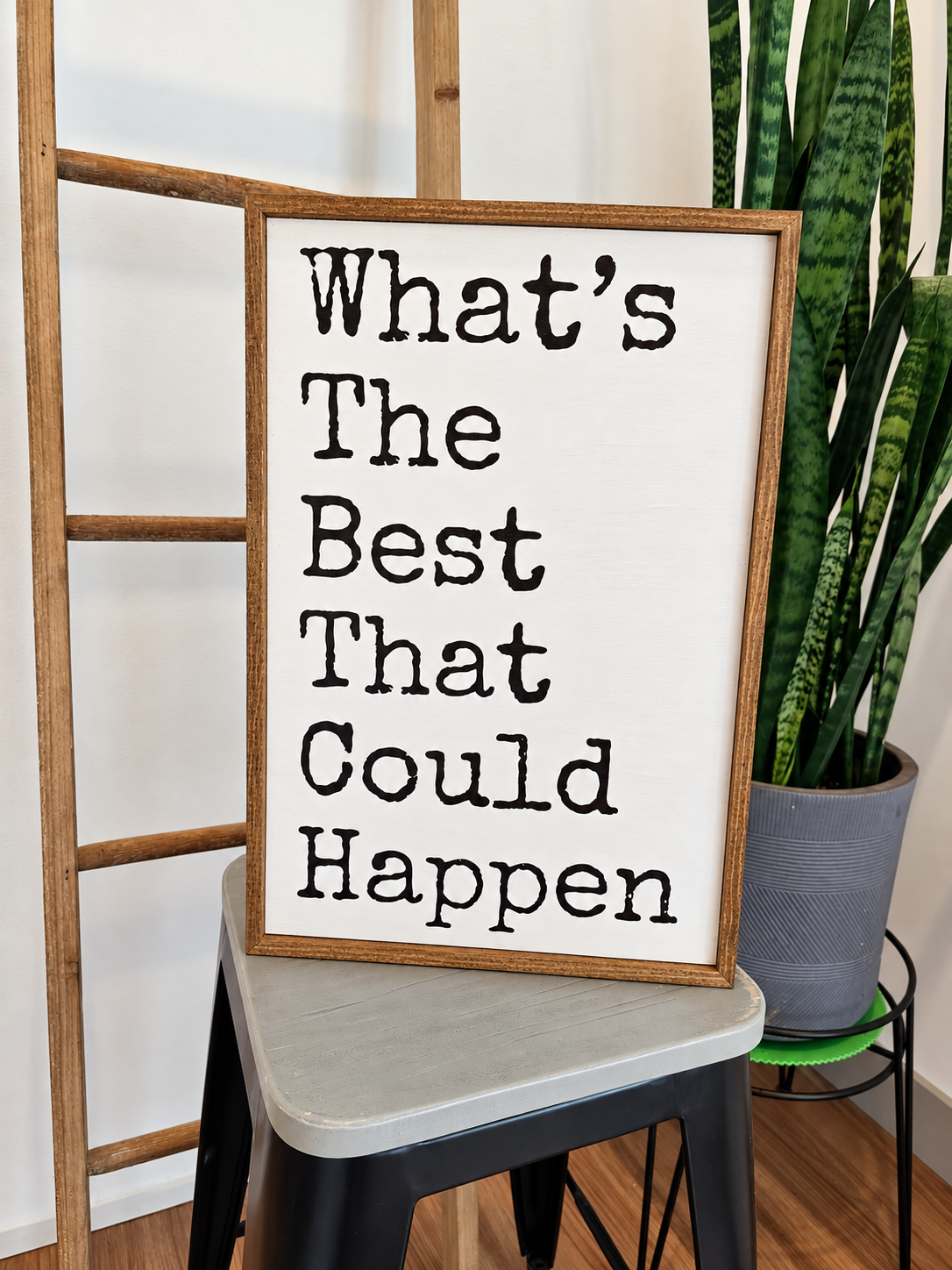 A framed typographic wall sign reading “What’s The Best That Could Happen” in black text on an off-white background, styled leaning on a stool in a home setting with a plant and wooden ladder visible.