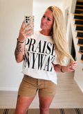 A woman taking a mirror selfie in a home interior, wearing a white graphic t-shirt with the text 'PRAISE ANYWAY' in black letters, paired with tan shorts.
