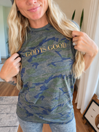 A woman wearing a heather gray and blue camouflage graphic t-shirt with 'GOD IS GOOD' printed in gold across the chest, posing indoors.