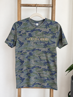A short-sleeve crewneck t-shirt with a blue and green camouflage pattern, featuring the text 'GOD IS GOOD' printed in gold across the chest. The shirt is displayed on a wooden hanger hanging from a rustic wooden ladder against a white wall.