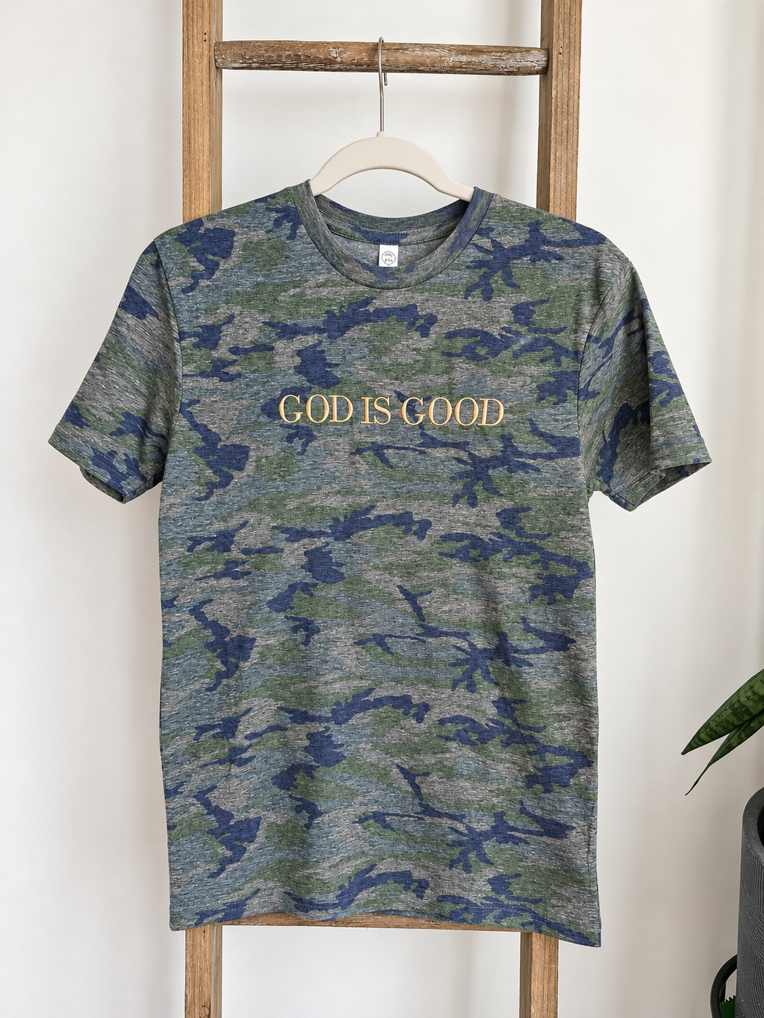 A short-sleeve crewneck t-shirt with a blue and green camouflage pattern, featuring the text 'GOD IS GOOD' printed in gold across the chest. The shirt is displayed on a wooden hanger hanging from a rustic wooden ladder against a white wall.
