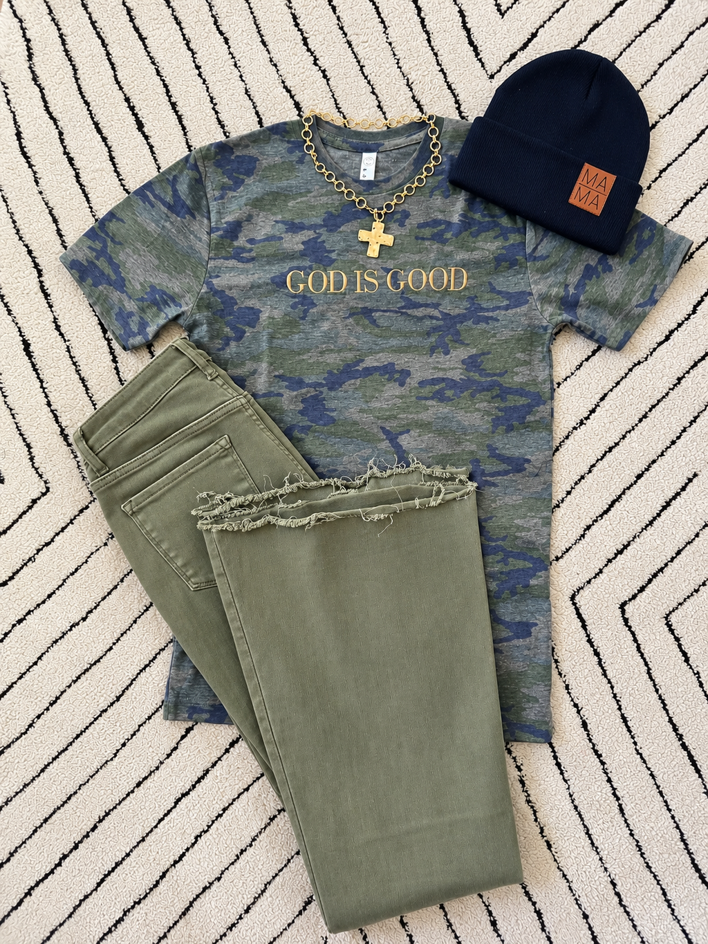 A styled flat-lay outfit featuring a blue and green camouflage graphic t-shirt with 'GOD IS GOOD' printed in gold, olive green frayed-hem jeans, a navy blue beanie with a brown 'MADE IN FLORIDA' patch, and a gold cross necklace, all arranged on a black and white striped rug.
