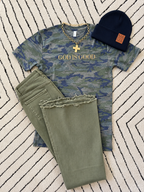 A styled flat-lay outfit featuring a blue and green camouflage graphic t-shirt with 'GOD IS GOOD' printed in gold, olive green frayed-hem jeans, a navy blue beanie with a brown 'MADE IN FLORIDA' patch, and a gold cross necklace, all arranged on a black and white striped rug.