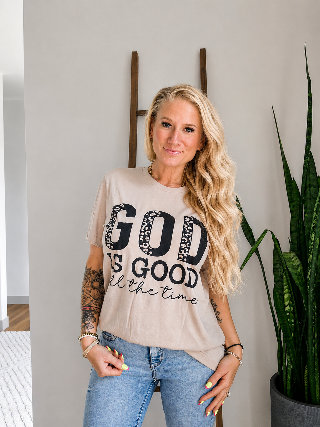 A woman with long blonde hair stands indoors wearing a beige graphic T-shirt with the text 'GOD IS GOOD all the time' in black, paired with blue jeans. She is posed in front of a light gray wall with a wooden ladder-style decor piece and a green potted snake plant.