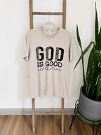 A beige graphic t-shirt hanging on a wooden ladder, featuring the text 'GOD IS GOOD all the time' in black. The scene is styled with a potted snake plant on a stand against a white wall and wood floor.