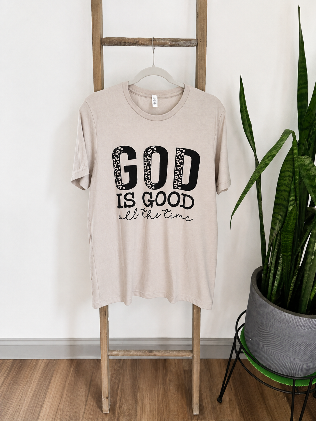 A beige graphic t-shirt hanging on a wooden ladder, featuring the text 'GOD IS GOOD all the time' in black. The scene is styled with a potted snake plant on a stand against a white wall and wood floor.