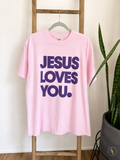 A light pink short-sleeve t-shirt hanging on a wooden hanger from a wooden ladder, featuring the text 'JESUS LOVES YOU.' printed in dark purple.
