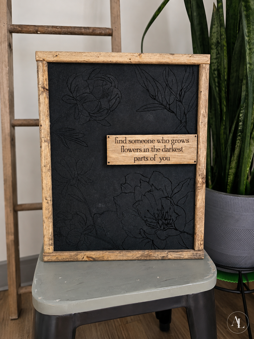 A rustic framed black chalkboard sign with a weathered wood border and a small wooden plaque that reads, 'find someone who grows flowers in the darkest parts of you.' The sign is displayed on a stool in an indoor setting next to a potted snake plant.