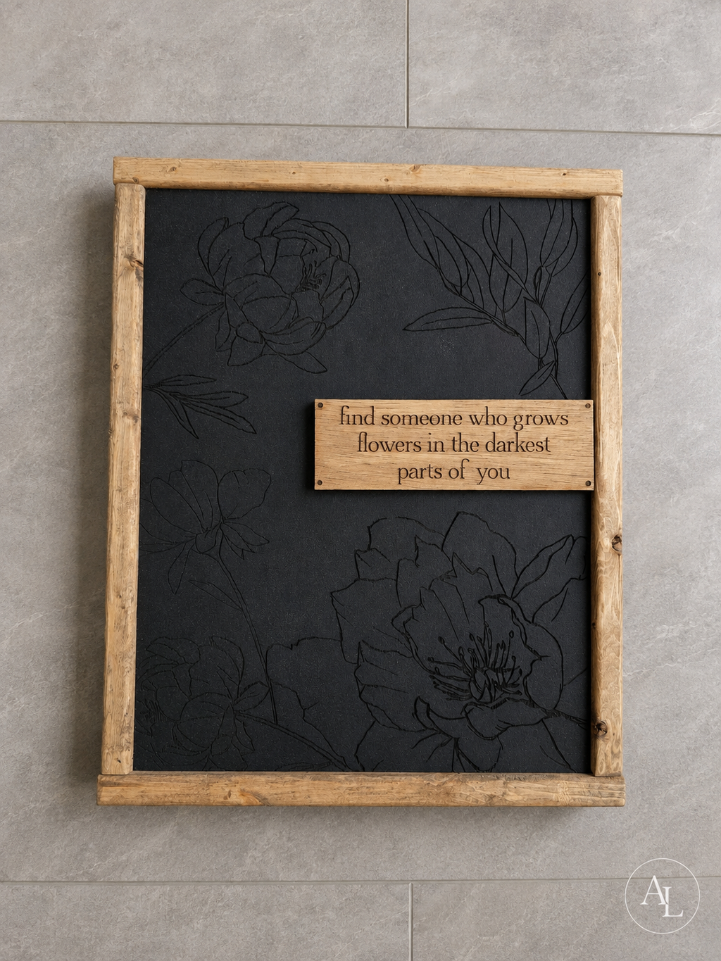 A square framed wall art piece with a dark charcoal background, subtle floral line art, and a central wooden plaque that reads, 'find someone who grows flowers in the darkest parts of you.' The frame is a natural light wood tone, and the piece is photographed against a light gray tiled wall.