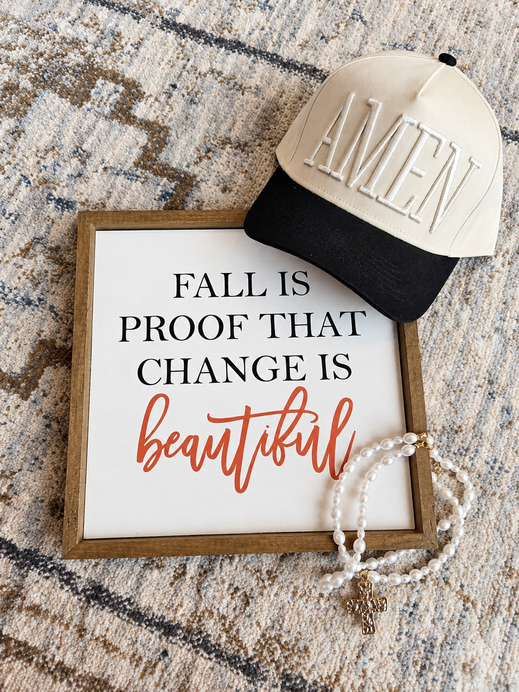 A styled flat-lay featuring a square framed sign with the text 'FALL IS PROOF THAT CHANGE IS beautiful' in black and orange lettering on a white background. A cream and black trucker hat with 'AMEN' embroidered on the front rests on top, and a white beaded necklace with a gold cross is draped over the frame.