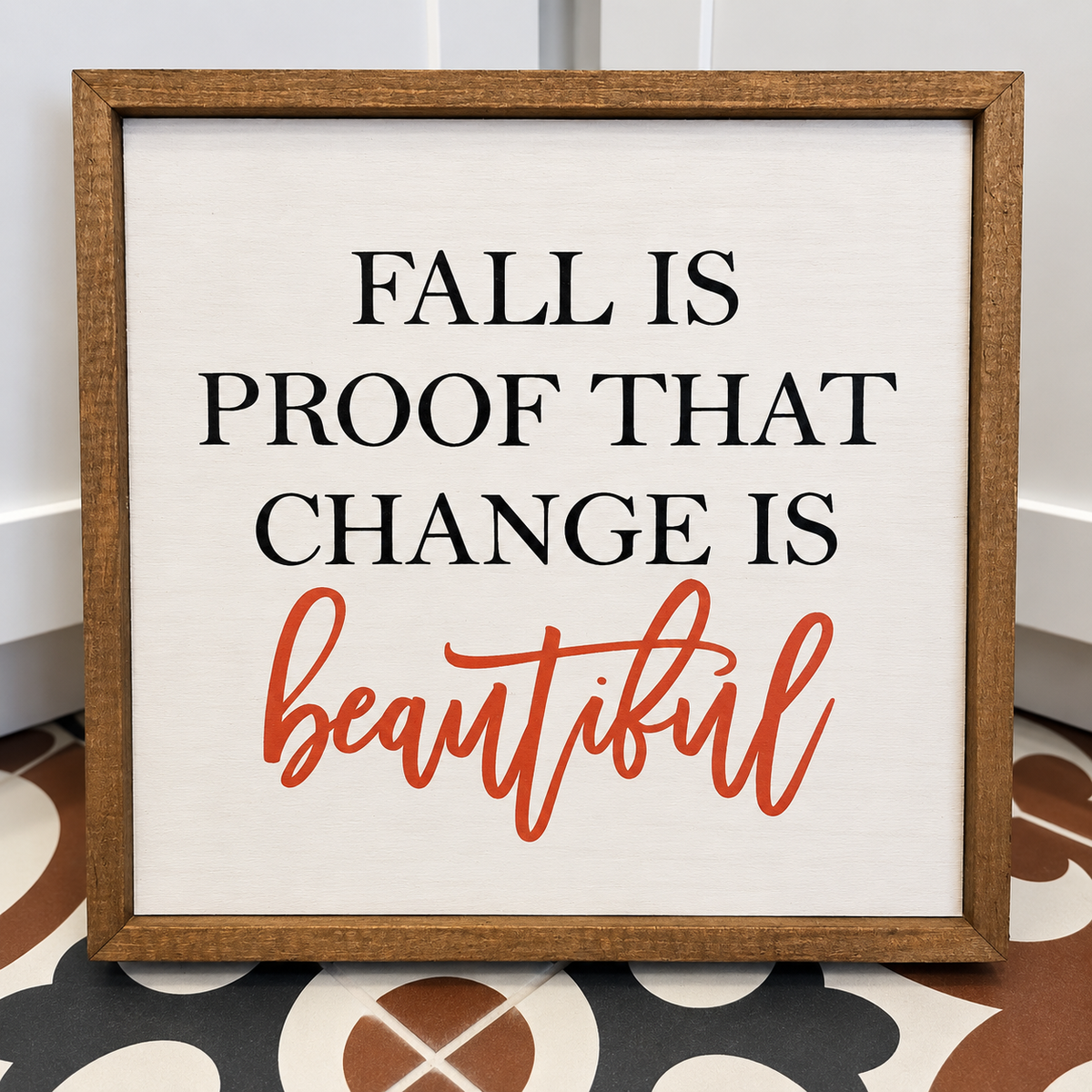 A square framed decorative sign with the text 'FALL IS PROOF THAT CHANGE IS beautiful' in black and orange lettering on a white background, set in a rustic brown wood frame.