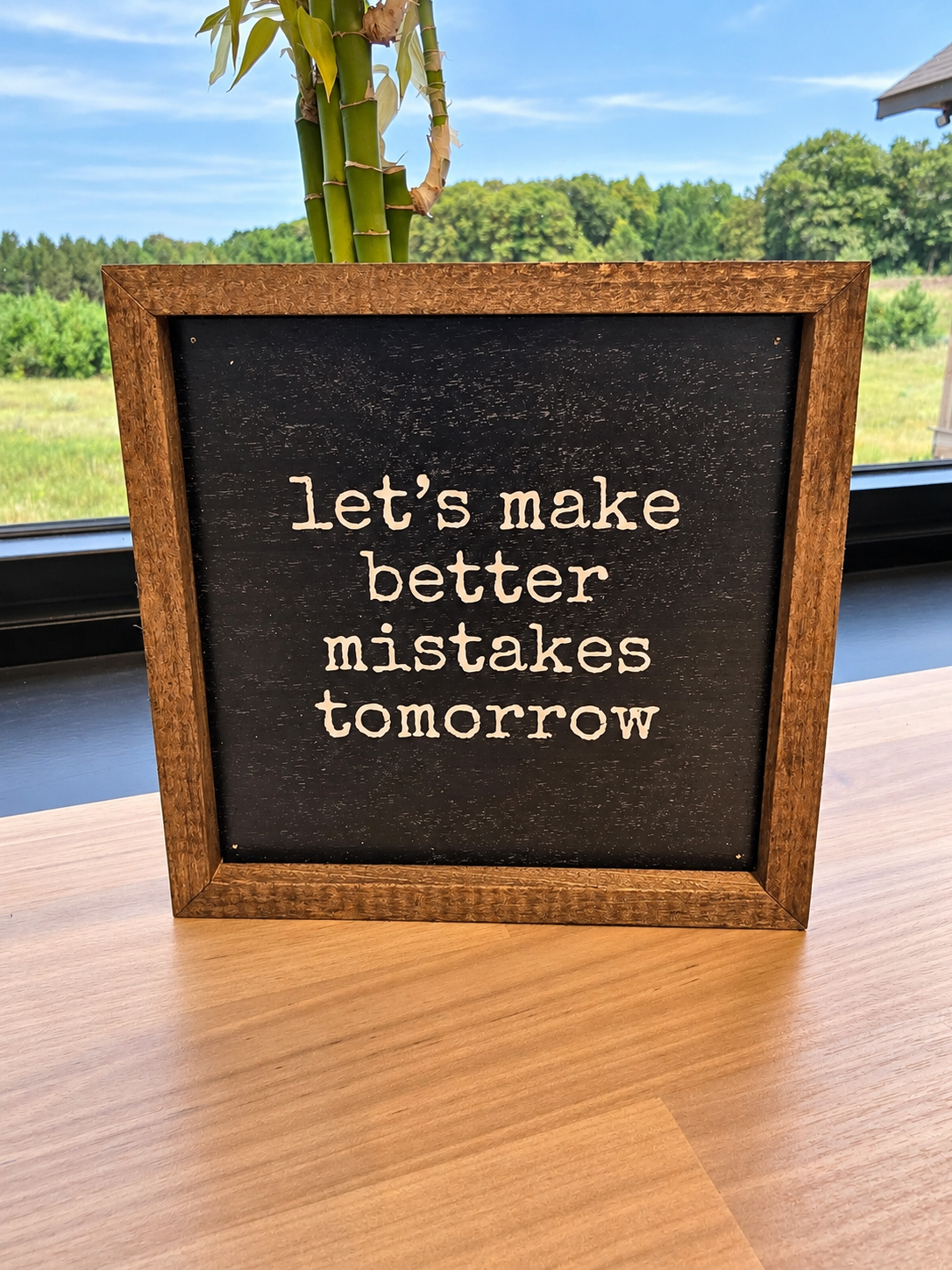 A square framed sign with a dark chalkboard-style center and light beige text reading “let’s make better mistakes tomorrow,” set on a wooden table with a window and outdoor landscape in the background.