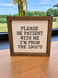 A square wooden-framed sign with black lettering on a cream background, reading 'PLEASE BE PATIENT WITH ME I'M FROM THE 1900's', displayed on a wooden table with a window and bamboo plant in the background.