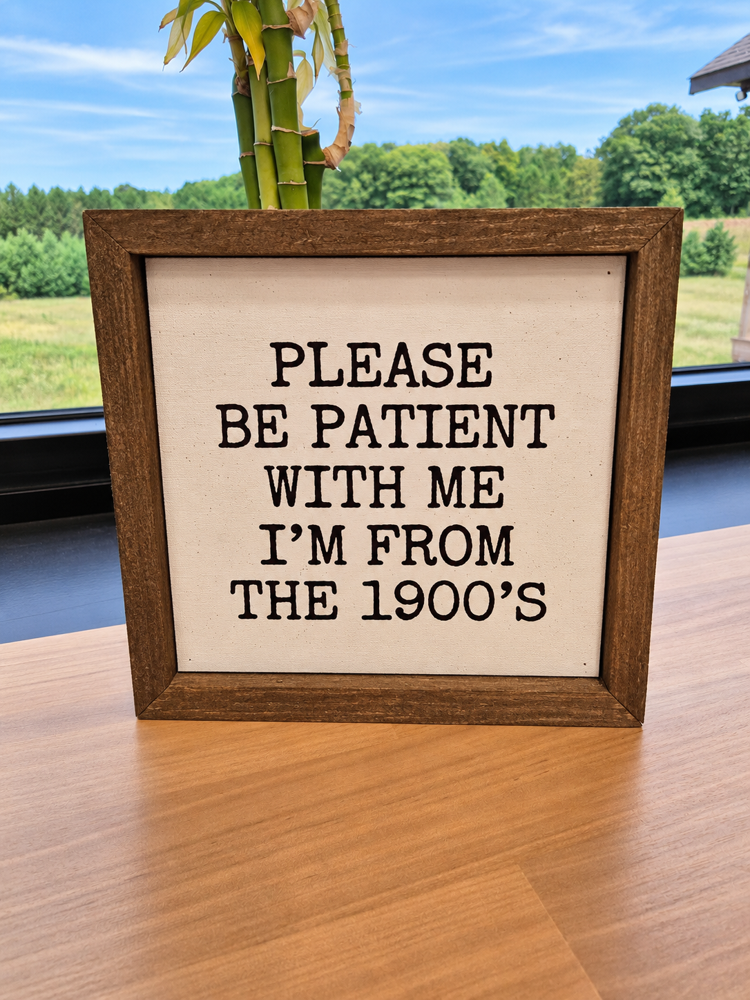 A square wooden-framed sign with black lettering on a cream background, reading 'PLEASE BE PATIENT WITH ME I'M FROM THE 1900's', displayed on a wooden table with a window and bamboo plant in the background.