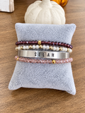 Four beaded and metal bracelets displayed on a light blue plush jewelry pillow over a wooden surface. The bracelets include dark burgundy beads, white faux pearls, a silver cuff stamped with the name 'SELAH', and light pink faceted beads, each with small gold spacer beads.
