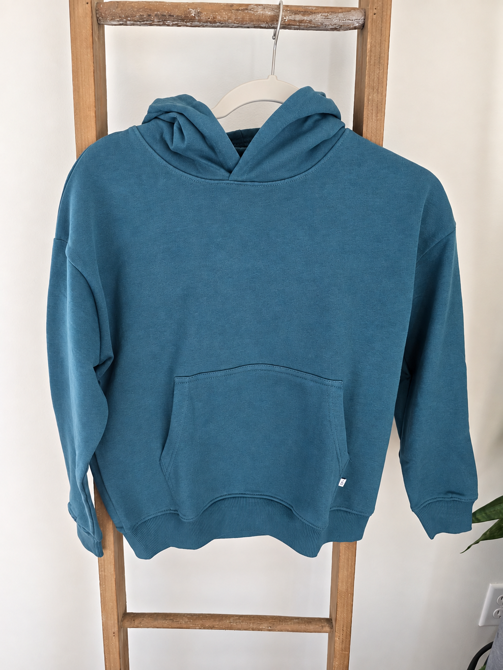 A teal blue hooded sweatshirt with a front kangaroo pocket, hanging on a white hanger from a wooden ladder against a white wall.