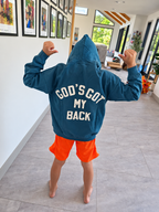 A child standing with their back to the camera in a modern home interior, wearing a teal blue hoodie with 'GOD'S GOT MY BACK' printed in white on the back, paired with bright orange shorts.