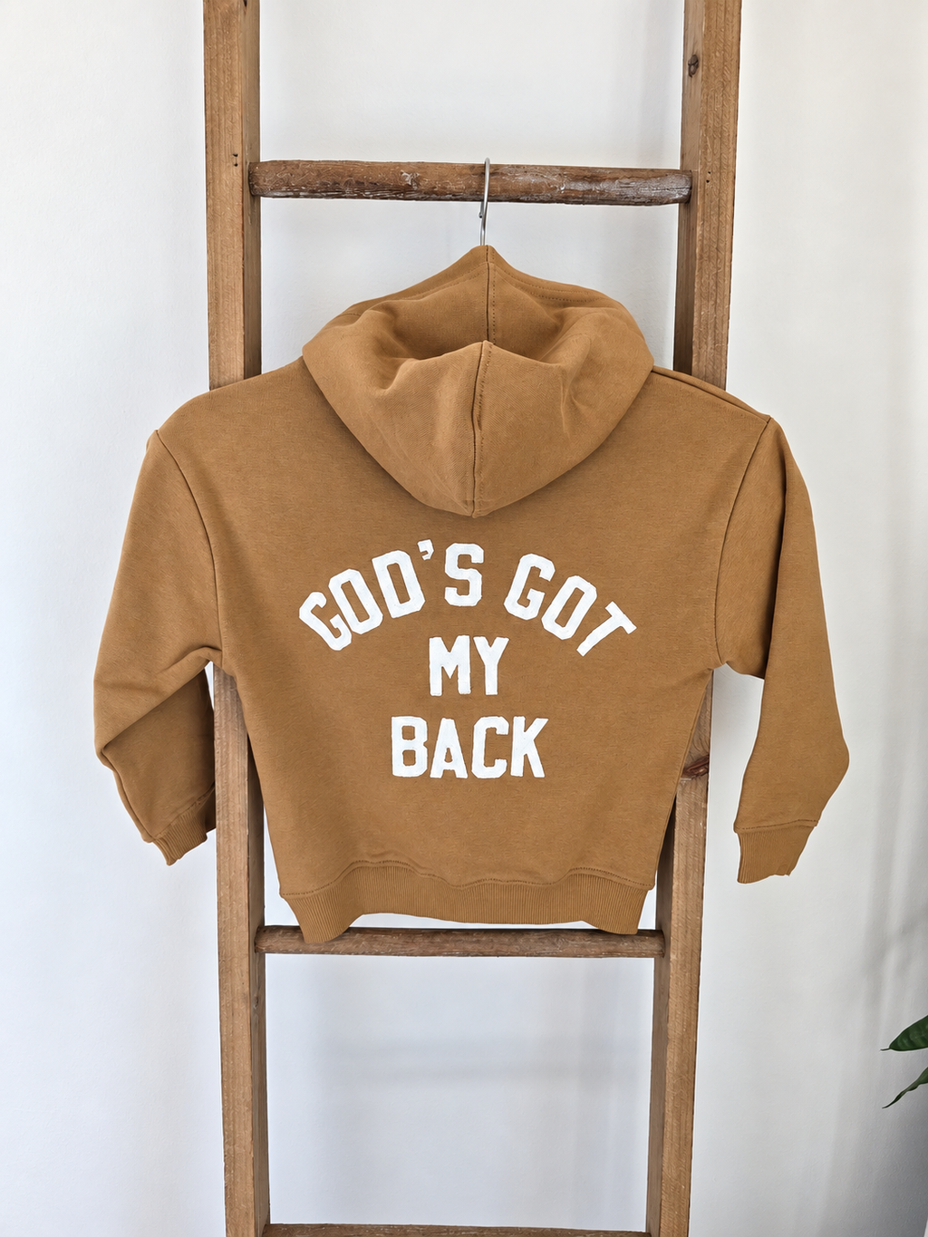A tan hoodie with white text reading 'GOD'S GOT MY BACK' on the back, hanging on a wooden ladder against a white wall.