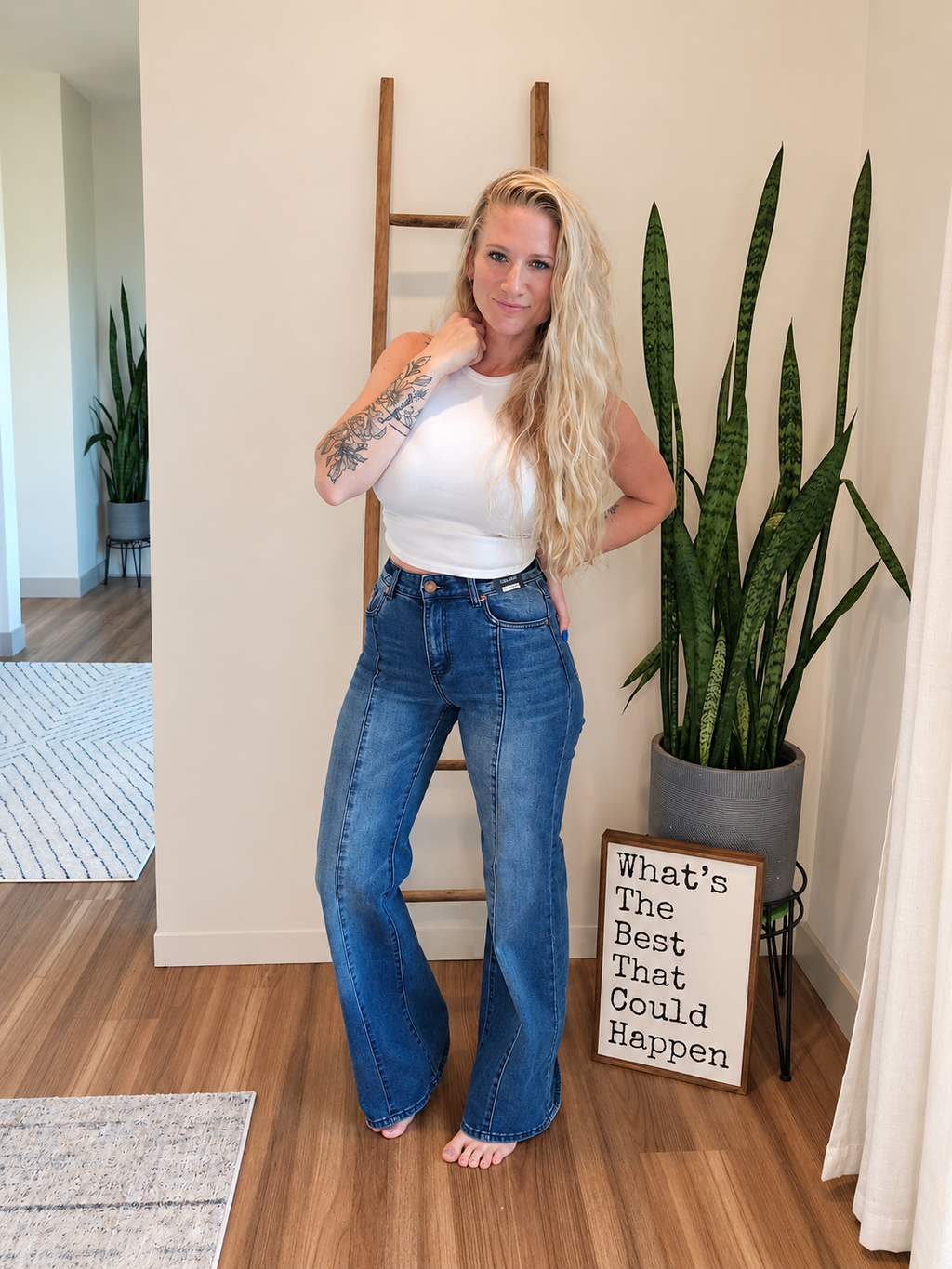 A woman with blonde hair stands barefoot in a home interior wearing a white tank top and high-waisted, wide-leg blue jeans. She is posing next to a wooden ladder, a large potted snake plant, and a decorative sign that reads 'What's The Best That Could Happen'.