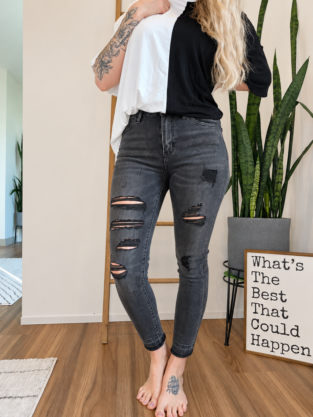 A model wearing distressed black skinny jeans with ripped knees, styled with a black t-shirt and white t-shirt, standing in a room with a wooden floor, potted plants, and a decorative sign.