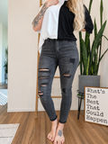 A model wearing distressed black skinny jeans with ripped knees, styled with a black t-shirt and white t-shirt, standing in a room with a wooden floor, potted plants, and a decorative sign.