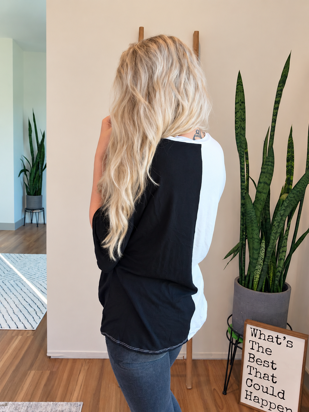 A woman with long blonde hair is shown from behind wearing a black and white raglan-style top and blue jeans in a modern indoor room with plants and wood flooring.