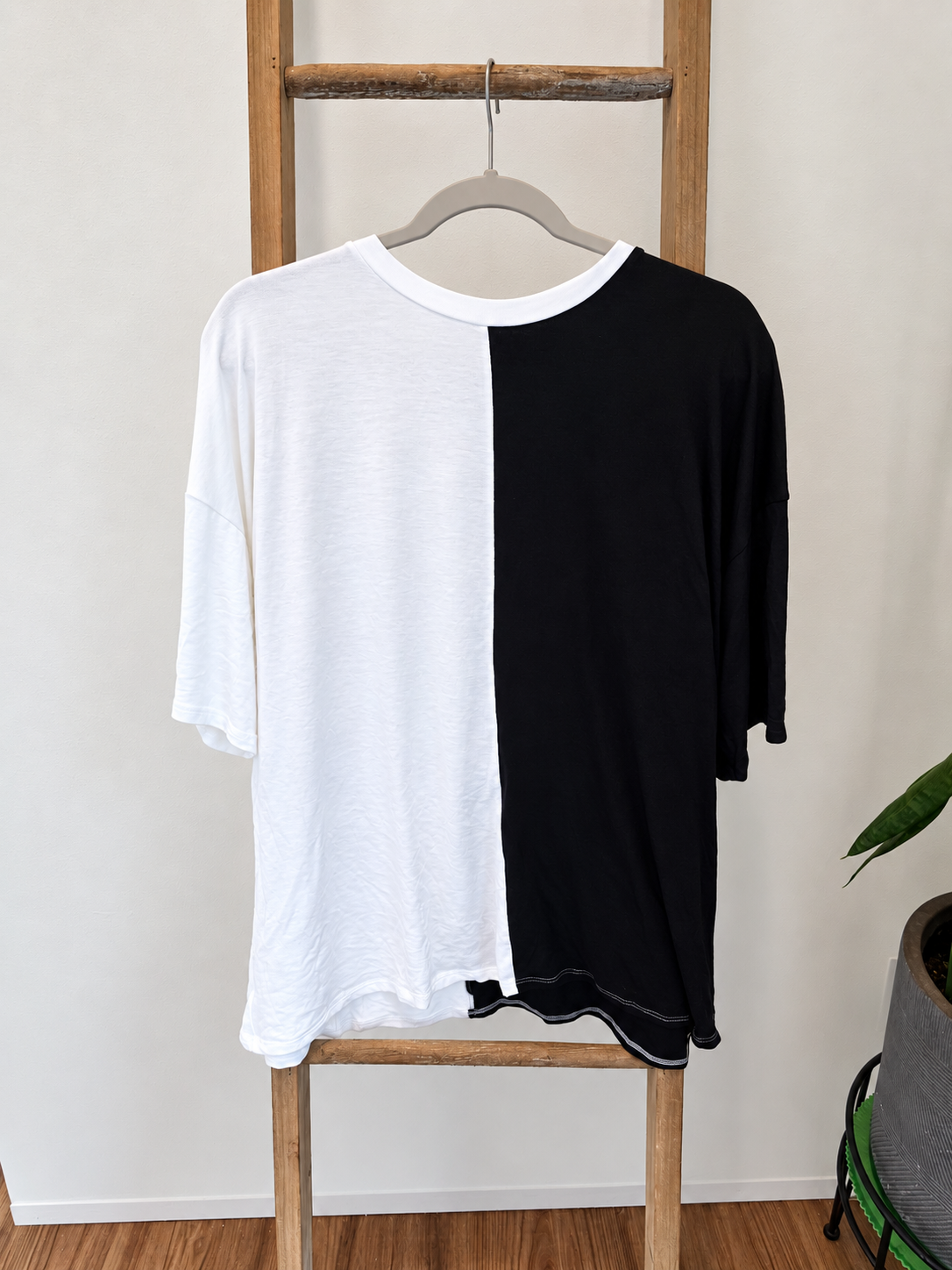 A split-color t-shirt with one half in white and the other in black, hanging on a wooden hanger on a wooden ladder against a white wall.