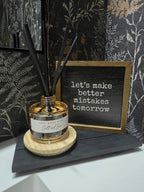 A reed diffuser bottle with black reeds sits on a round wooden coaster next to a small framed sign that reads 'let's make better mistakes tomorrow'. The scene is styled against dark botanical wallpaper.