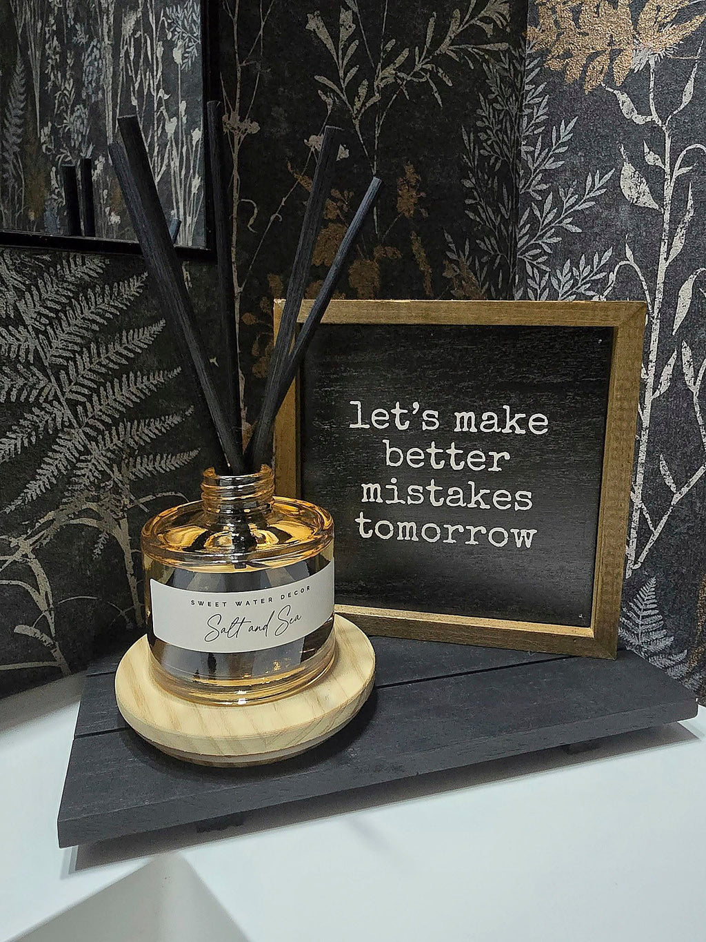 A reed diffuser bottle with black reeds sits on a round wooden coaster next to a small framed sign that reads 'let's make better mistakes tomorrow'. The scene is styled against dark botanical wallpaper.