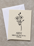 A cream-colored greeting card with a black floral illustration and the text 'ABIDE' and 'ABIDE IN ME, AND I IN YOU. JOHN 15:4' lies on top of a white envelope against a textured gray background.