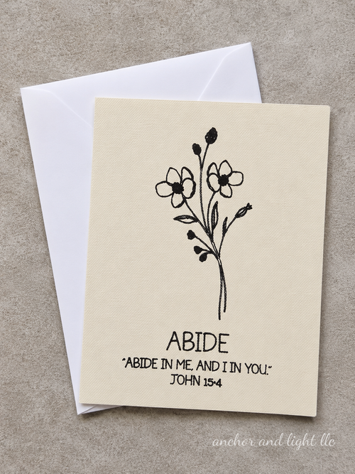 A cream-colored greeting card with a black floral illustration and the text 'ABIDE' and 'ABIDE IN ME, AND I IN YOU. JOHN 15:4' lies on top of a white envelope against a textured gray background.
