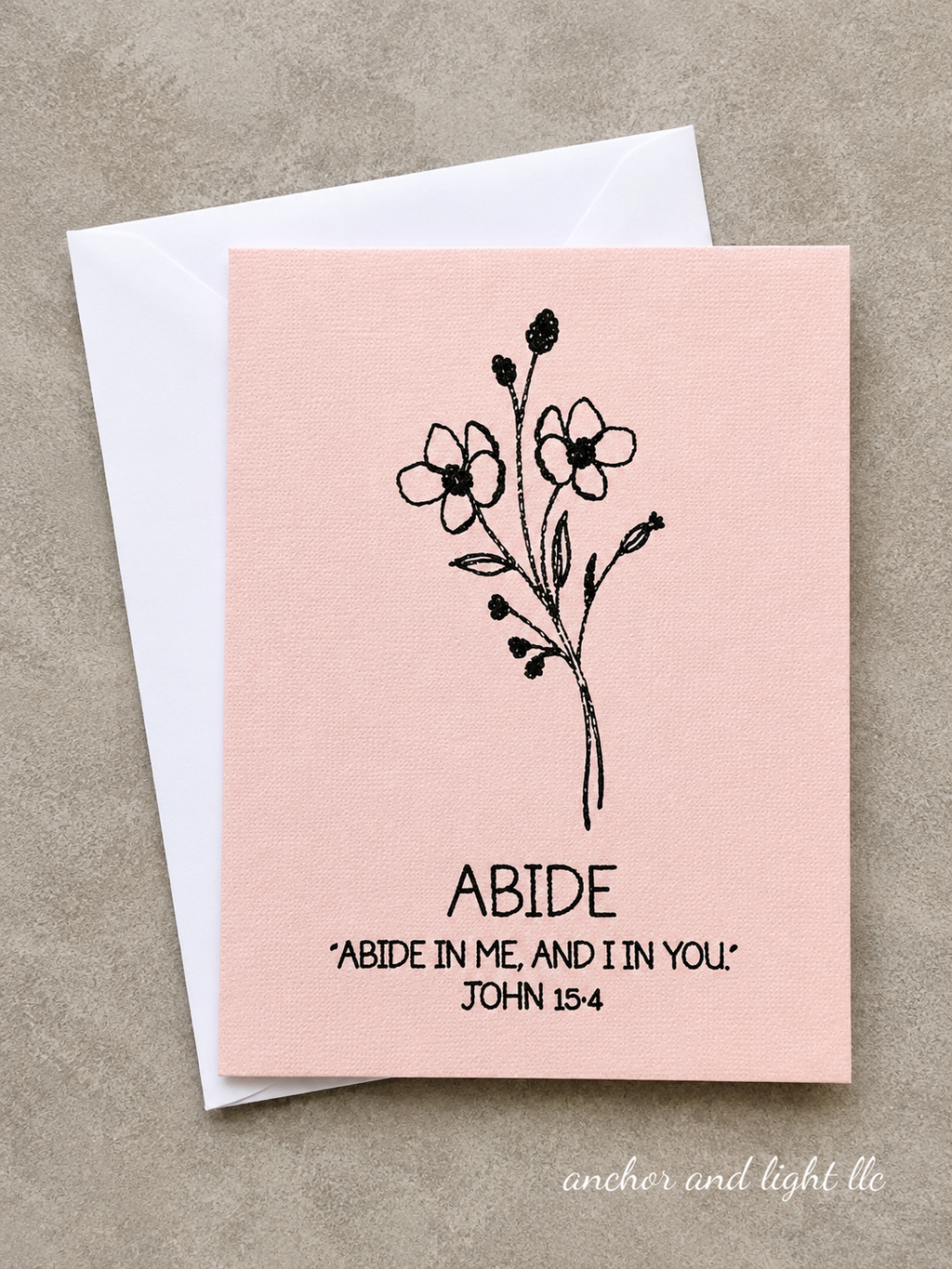 A square pink greeting card with a black floral illustration and the text 'ABIDE' and 'ABIDE IN ME, AND IN YOU. JOHN 15:4' printed on it, shown with a white envelope on a textured beige background.
