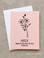 A square pink greeting card with a black floral illustration and the text 'ABIDE' and 'ABIDE IN ME, AND IN YOU. JOHN 15:4' printed on it, shown with a white envelope on a textured beige background.