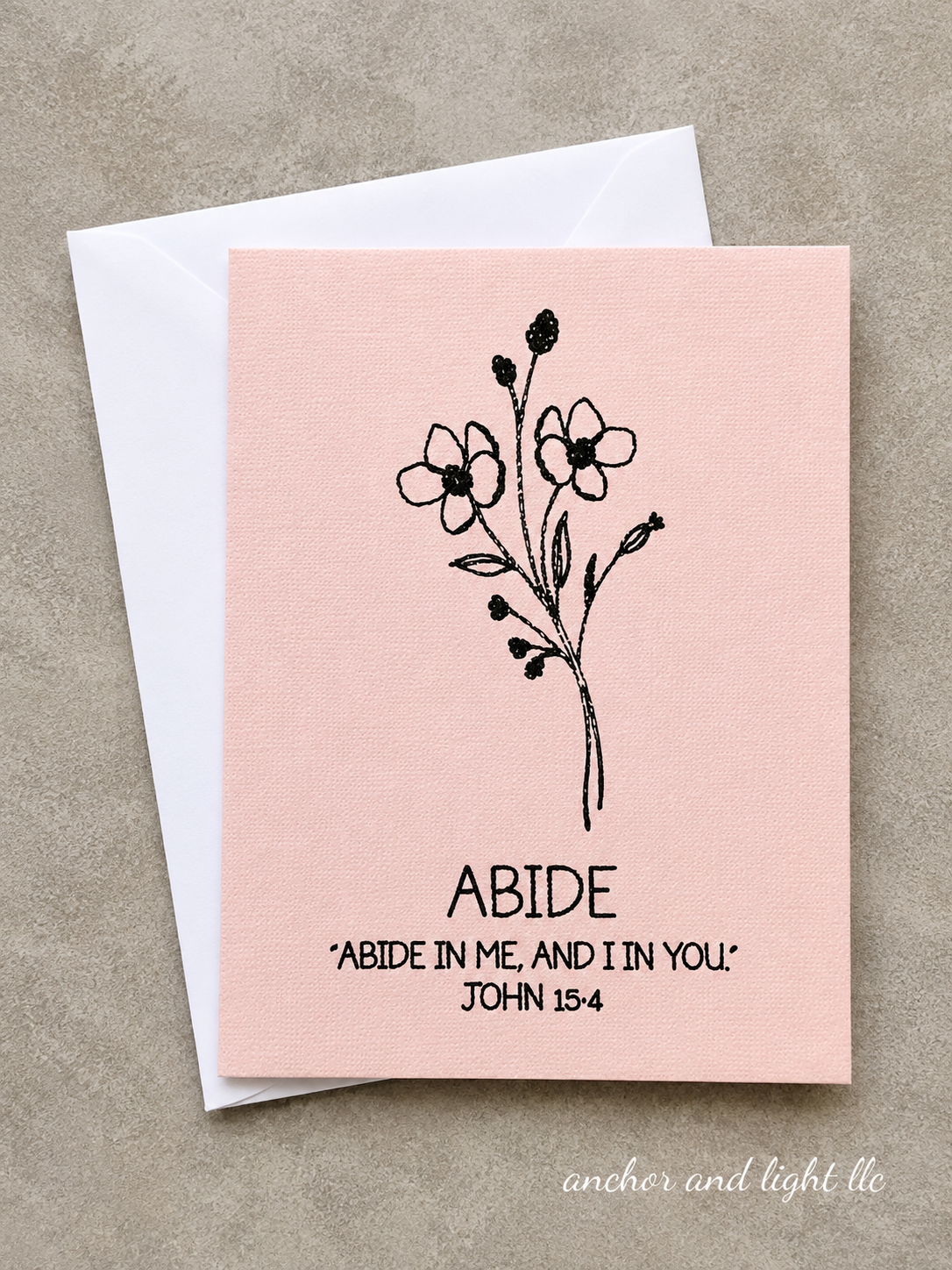 A square pink greeting card with a black floral illustration and the text 'ABIDE' and 'ABIDE IN ME, AND IN YOU. JOHN 15:4' printed on it, shown with a white envelope on a textured beige background.