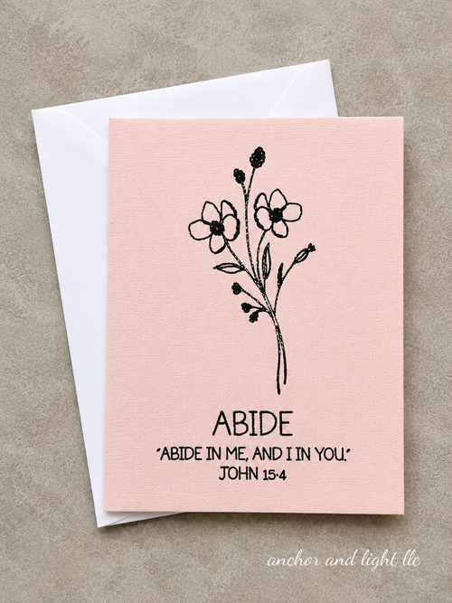 A square pink greeting card with a black floral illustration and the text 'ABIDE' and 'ABIDE IN ME, AND IN YOU. JOHN 15:4' printed on it, shown with a white envelope on a textured beige background.
