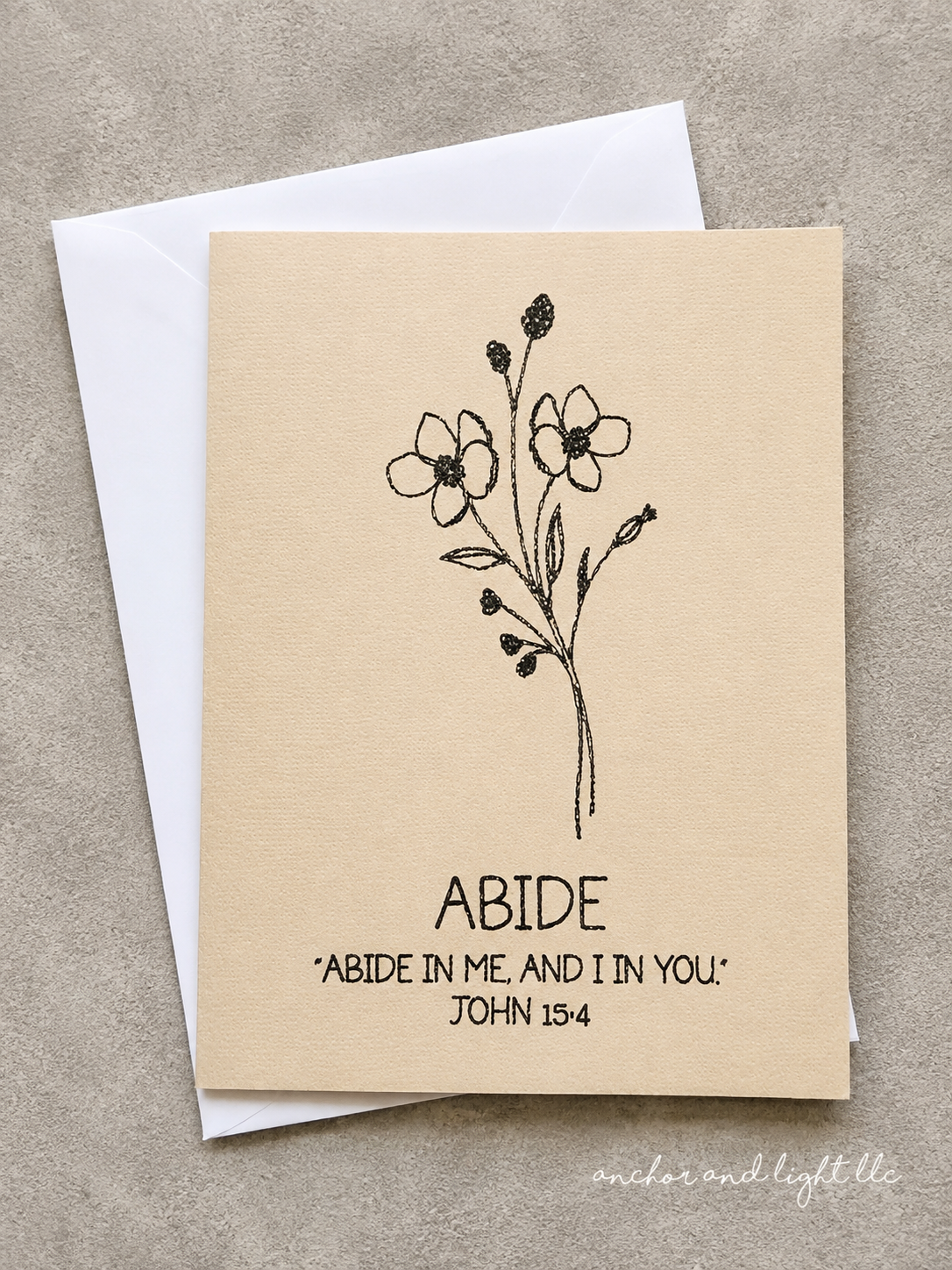 A square beige greeting card with a black floral line drawing and the printed text 'ABIDE' and 'ABIDE IN ME, AND I IN YOU' John 15:4, shown with a white envelope on a textured gray surface.
