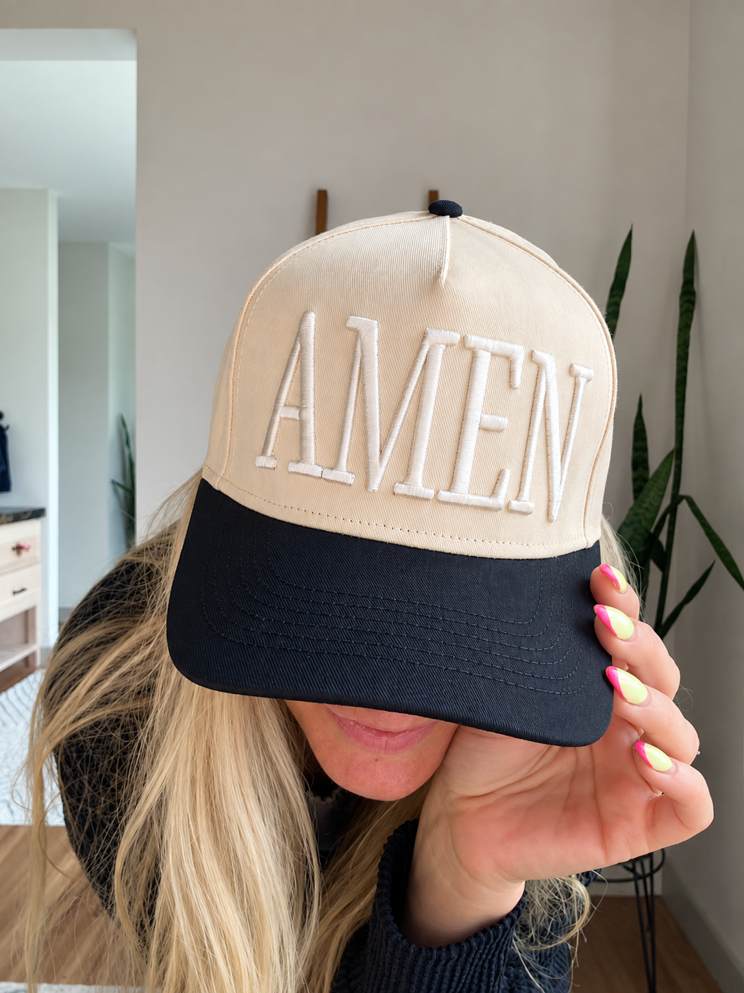 A person indoors wearing a beige and black baseball cap with the word 'AMEN' embroidered in white across the front, holding the brim over their eyes.