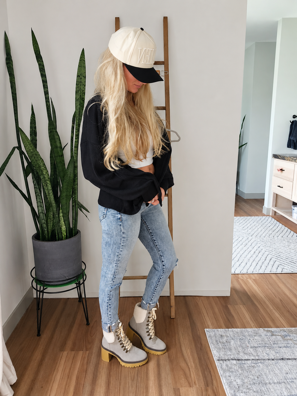 A woman stands in a room wearing a cream baseball cap with black trim, a black sweatshirt, light-wash blue jeans, and gray lace-up ankle boots with gold-toned hardware and yellow soles. She is posed next to a wooden ladder shelf and a potted snake plant.