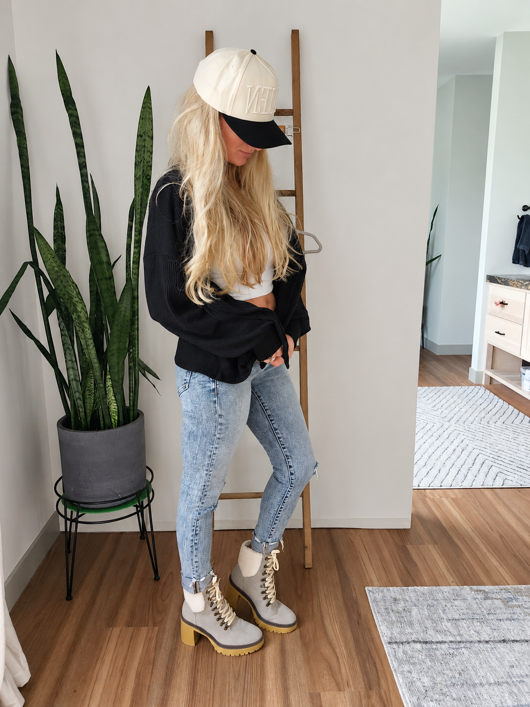 A woman stands in a room wearing a cream baseball cap with black trim, a black sweatshirt, light-wash blue jeans, and gray lace-up ankle boots with gold-toned hardware and yellow soles. She is posed next to a wooden ladder shelf and a potted snake plant.