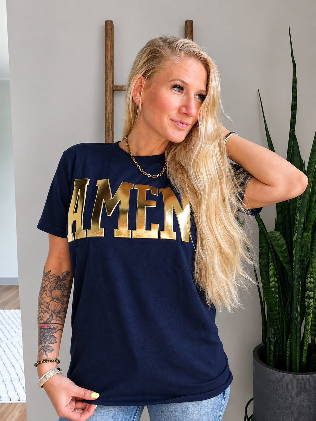 A blonde woman wearing a navy blue t-shirt with 'AMEN' printed across the chest in large gold letters, posed next to a potted snake plant in an indoor setting.