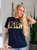 A blonde woman wearing a navy blue t-shirt with 'AMEN' printed across the chest in large gold letters, posed next to a potted snake plant in an indoor setting.