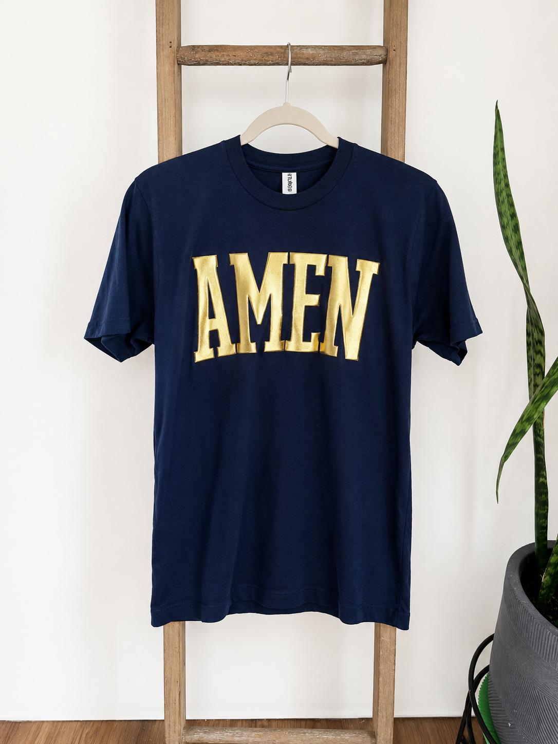 A navy blue short-sleeve t-shirt with 'AMEN' printed in large gold letters across the chest, hanging on a wooden hanger from a wooden ladder against a white wall, with a potted snake plant on the right.