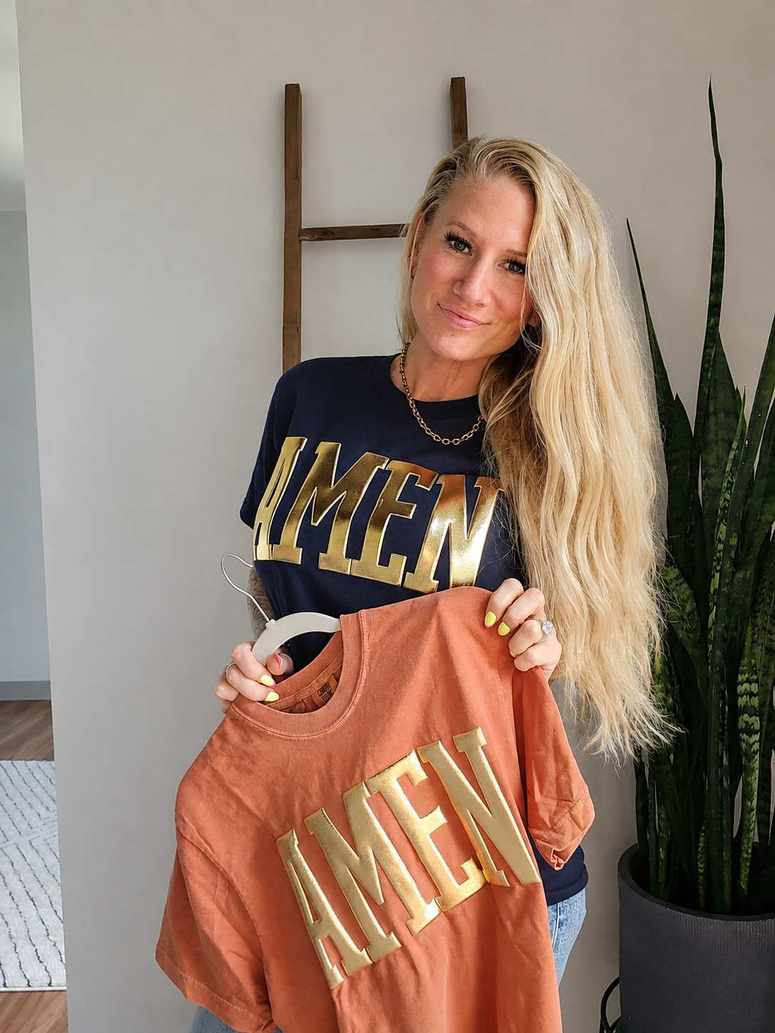 A woman with long blonde hair holds up a terracotta orange t-shirt on a hanger with gold lettering that says 'AMEN'. She is wearing a navy blue t-shirt with gold lettering that says 'MEN'.