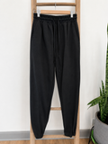 Black jogger sweatpants with an elastic waistband and drawstring, hanging on a wooden ladder against a white wall with a potted snake plant on the right.