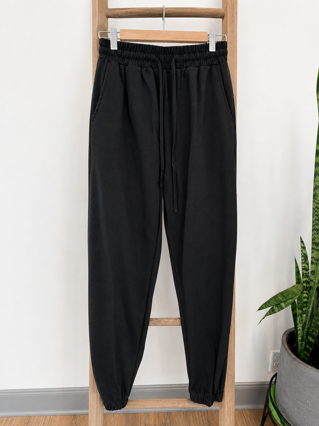 Black jogger sweatpants with an elastic waistband and drawstring, hanging on a wooden ladder against a white wall with a potted snake plant on the right.