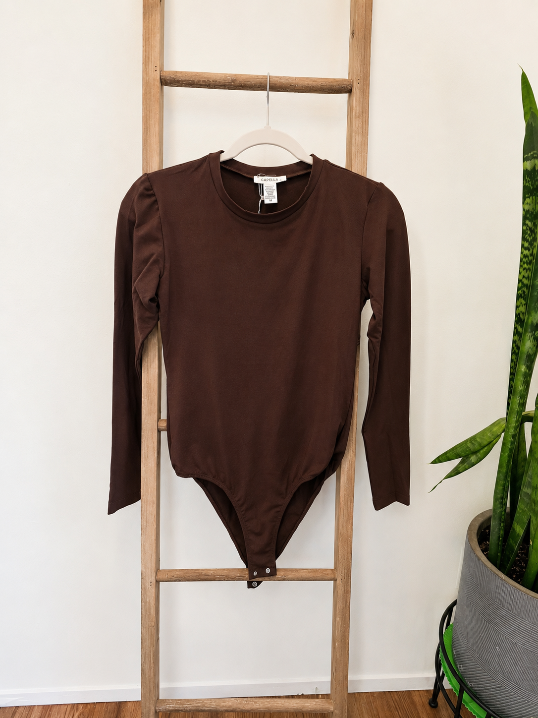 A dark brown long-sleeve bodysuit with puffed shoulders hangs on a wooden hanger from a wooden ladder, with a potted snake plant on the right against a white wall.