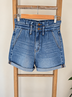 Light blue denim shorts with a high waist, pleated front, and rolled cuffs, hanging on a wooden hanger from a wooden ladder against a light beige wall.